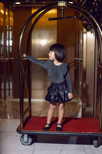 Beautiful korean girl child in a black dress is sitting on a luggage trolley in a hotel