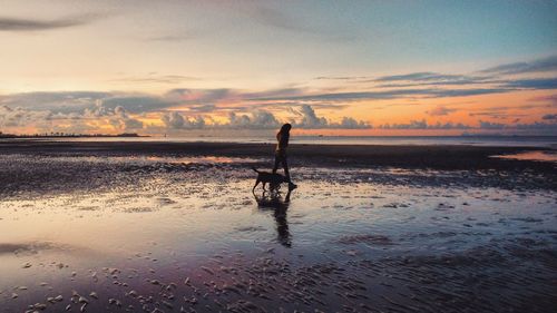 Woman walking on shore at beach against sky during sunset