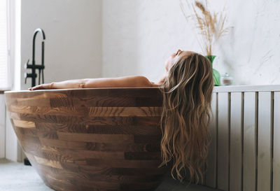 Blonde sensual young woman with long hair takes wooden bath relax at modern bathroom 