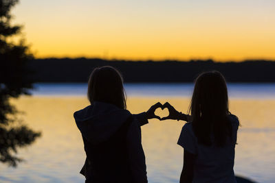 Rear view of girls forming heart shape with hands against lake during sunset