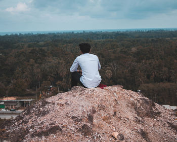 Rear view of man looking at land while sitting on rock