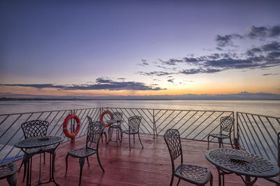 Empty chairs and table by sea against sky during sunset