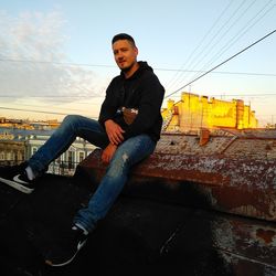 Young man looking away while sitting against sky during sunset