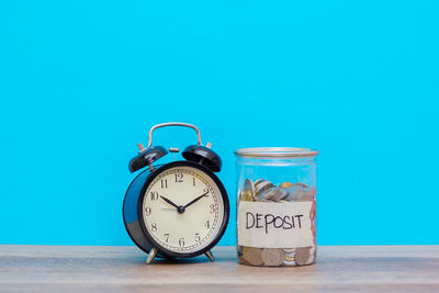 Close-up of alarm clock and coins in jar on table against blue background