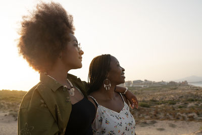 Happy young african american female friends in stylish summer outfits and accessories hugging and looking away while resting together on seashore in summer evening