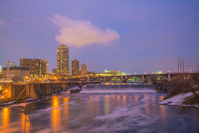 Illuminated buildings by river against sky in city