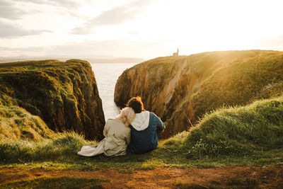 Back view of anonymous man and woman in outerwear leaning on each other and observing sea while sitting on cliff at sundown in aviles, spain