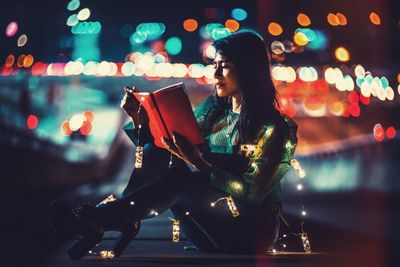 Side view of woman reading books at night near a busy road