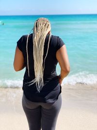 Rear view of young woman with dreadlocks standing at beach against sky during sunny day