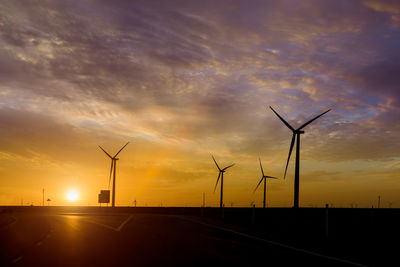 Silhouette wind turbines on road against sky during sunset