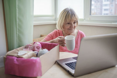 Senior woman holding coffee cup using laptop at home