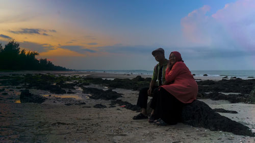 Couple sitting on beach against sky during sunset