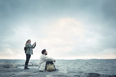 Rear view of friends sitting on sea shore against sky