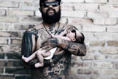 Portrait of tattooed father holding cute baby girl while standing against brick wall