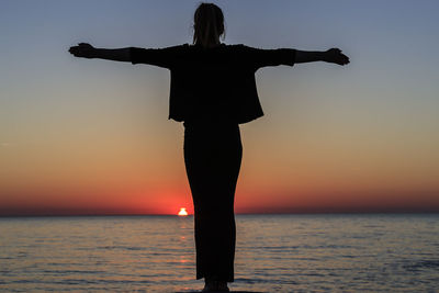 Silhouette woman with arms outstretched standing by sea against sky during sunset