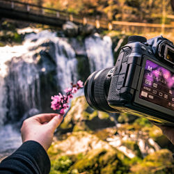 Close-up of hand holding camera against blurred waterfall and bridge in background
