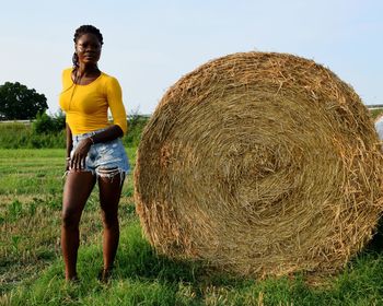 Full length portrait of woman standing by hay bale on field