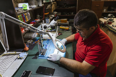 Worker looking at broken smart phone through magnifying glass in repair shop