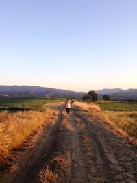 Rear view of woman walking on dirt road amidst field against sky