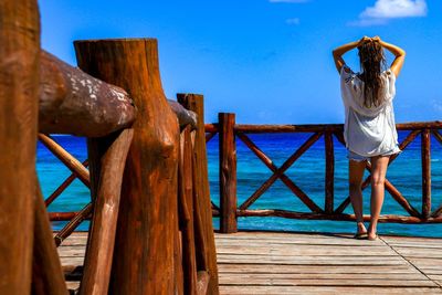 Rear view of young woman with hand in hair standing on pier over sea against blue sky