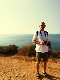 Portrait of man standing with camera against clear sky on sunny day