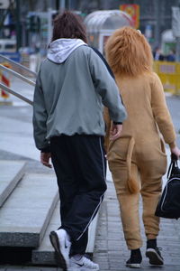 Rear view of person in lion costume walking with man on sidewalk