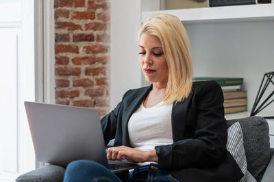 Cheerful blonde businesswoman smiling and browsing laptop while sitting in comfortable armchair near fireplace in cozy room working remotely from home