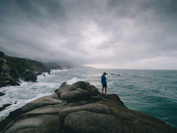 Rear view of man standing on rock by sea against cloudy sky