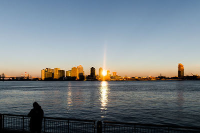 Silhouette city by river against clear sky during sunset