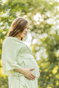 Young pregnant woman holds big belly, green trees,sky on background. brown long haired female
