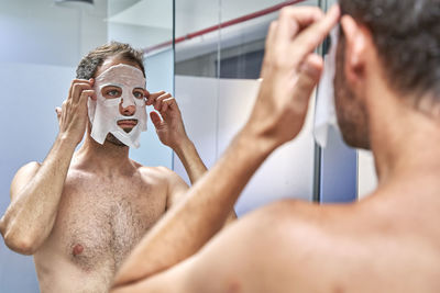 Man applying sheet mask on face in front of mirror