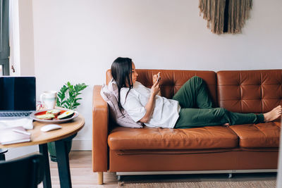 Portrait of young brunette female texting on smartphone while chilling on sofa at home.