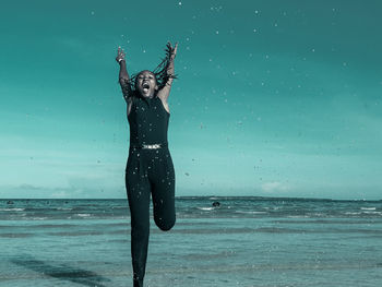 Full length of young woman with arms raised jumping in sea against sky