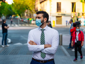 Young elegant businessman stands in middle of crowded street with surgical mask