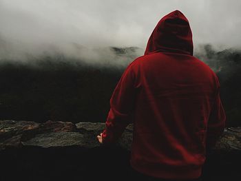 Rear view of man in red hooded shirt looking at mountains during foggy weather