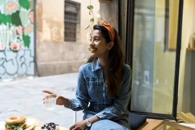 Smiling young woman eating food while sitting by window at cafe