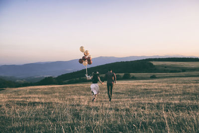 Young couple running with balloons on field against sky during sunset
