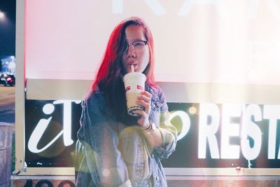 Young woman drinking while standing against billboard at night