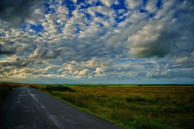 Road passing through field against cloudy sky