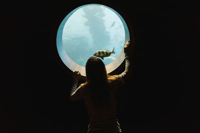 Back view of anonymous woman in casual wear looking at rounded window of aquarium with ocean tropical fishes