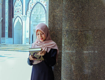 Young woman holding smart phone while standing against wall