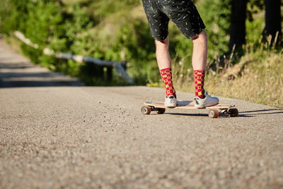 Crop of male hipster in fancy clothes riding skateboard along road in park in summer