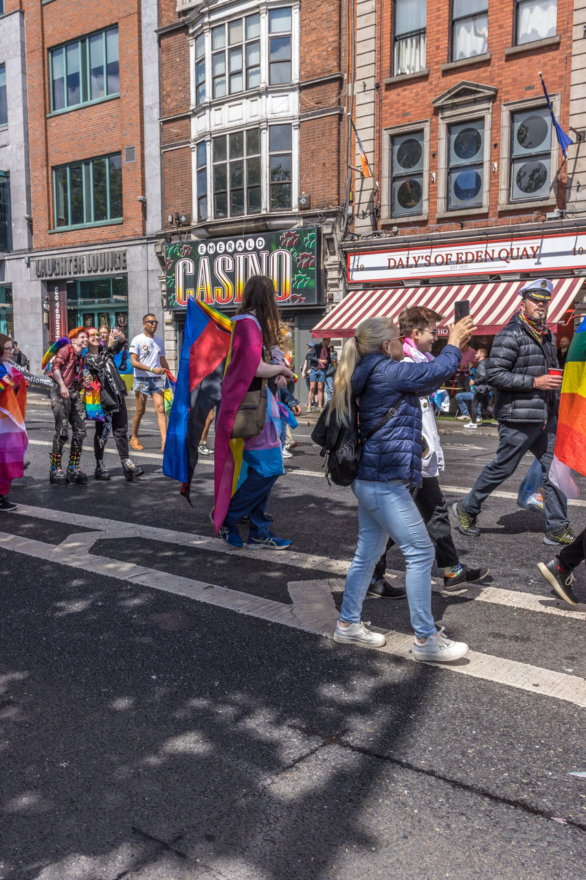 city, architecture, building exterior, group of people, street, built structure, crowd, men, day, women, large group of people, road, adult, city life, festival, parade, city street, walking, celebration, lifestyles, full length, outdoors, transportation, leisure activity, person, clothing, sunlight, event