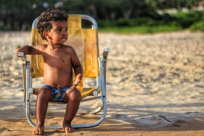 Full length of shirtless baby boy sitting on chair at beach
