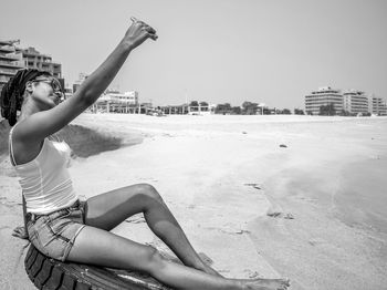 Young woman taking selfie from mobile phone while sitting on spare tire at sandy beach