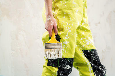 Cropped image of woman wearing dirty coveralls holding paintbrush while standing against wall