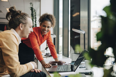 Smiling businesswoman leaning on desk while looking at businessman sitting at office