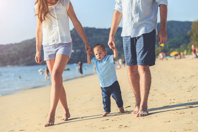 Low section of mother and father walking with son on sand at beach