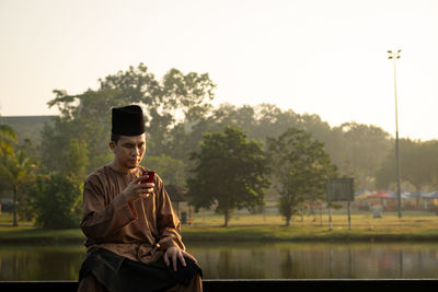 Man wearing traditional clothing using mobile phone while sitting on railing against lake during sunset