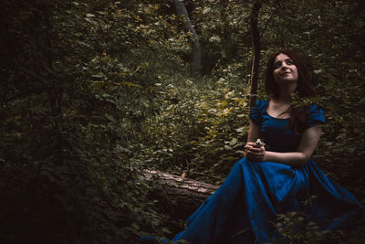 Thoughtful woman in blue dress looking up while sitting on tree trunk at forest